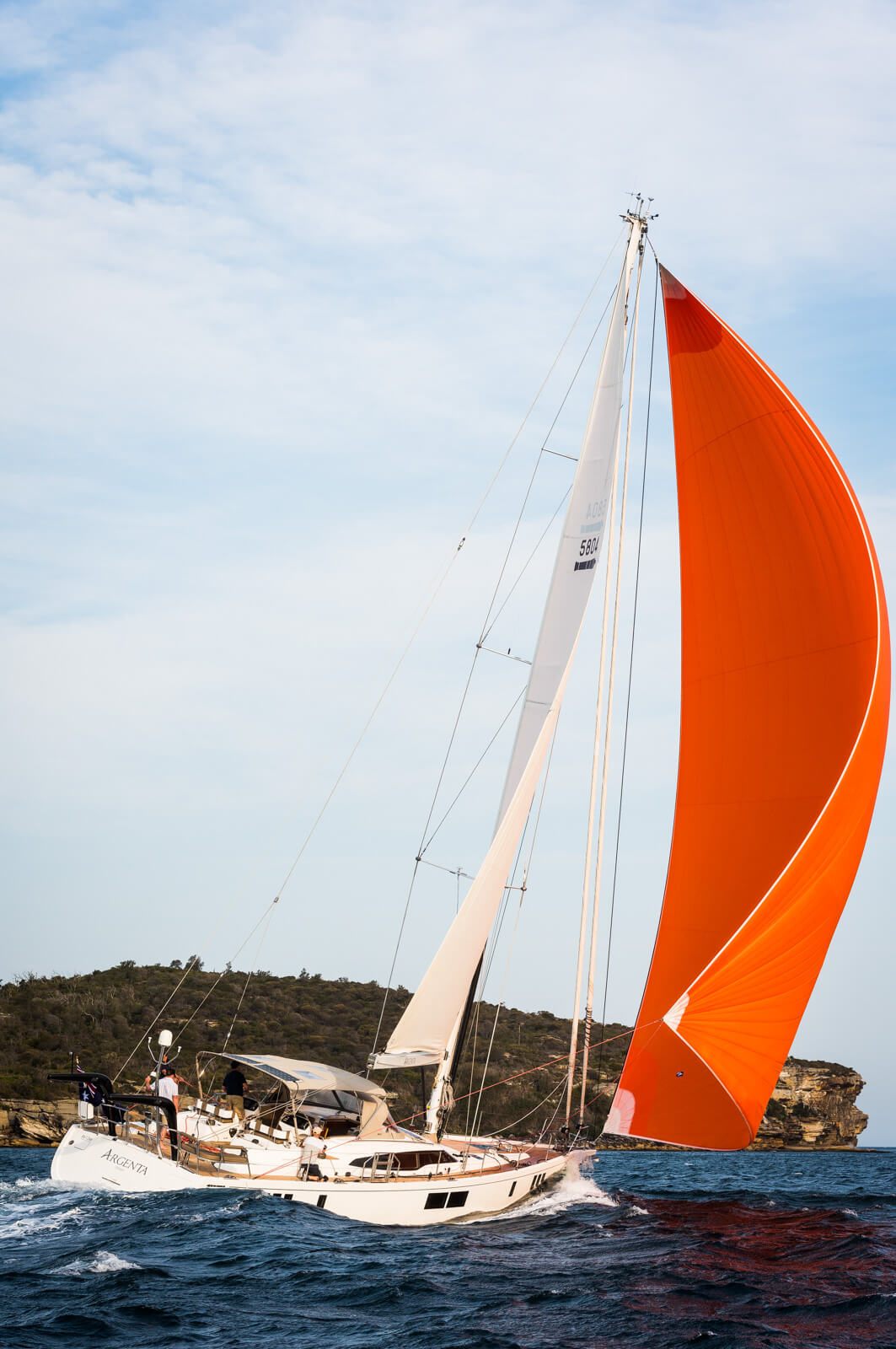 A white sailboat with a large orange sail glides across blue water near a rocky, green shoreline under a partly cloudy sky.