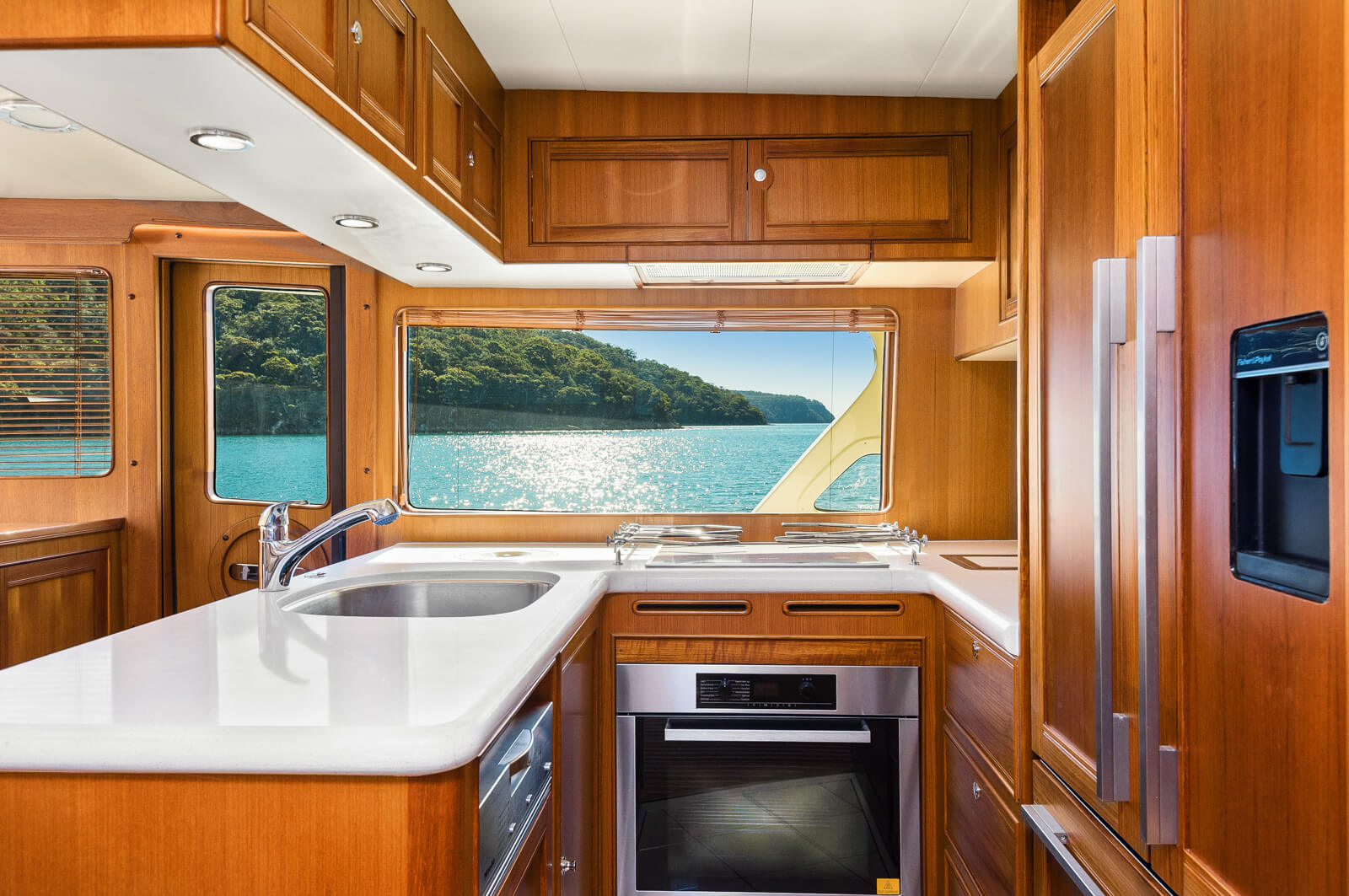 A modern yacht kitchen with wood cabinets, white countertops, a built-in oven, and a large window overlooking blue water and a lush green shoreline in bright sunlight.